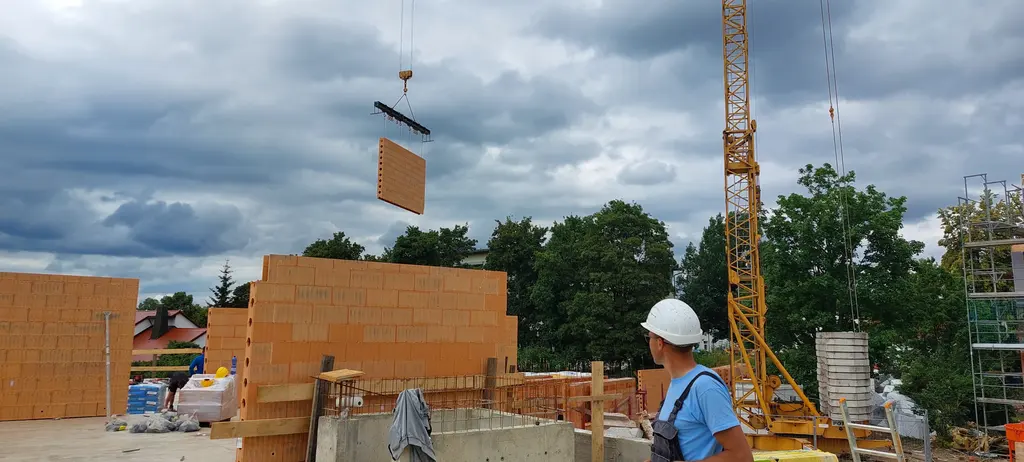 Ziegel-Wandelement am Turmdrehkran über einer Mauerwerks-Modulbaustelle, davor ein Arbeiter mit Helm
