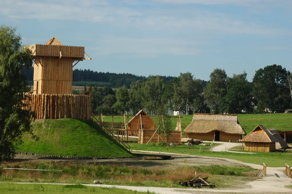 Holzturm der Motte auf grünem Erdhügel mit Palisade, dahinter Holzhäuser des Dorfs im Geschichtspark Bärnau-Tachov